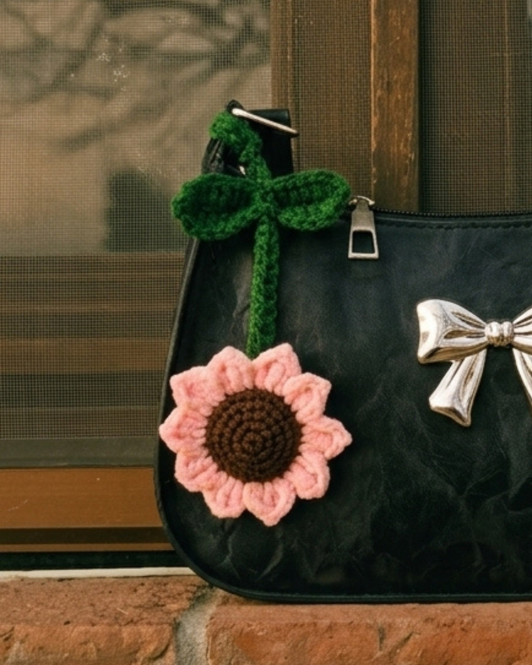 A close-up of a black handbag with a crocheted pink sunflower charm hanging from a green strap, set against a rustic window and brick background.