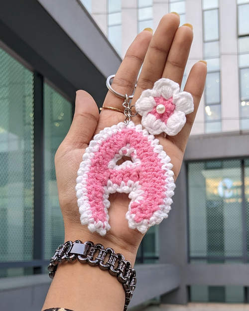 A hand holds a crocheted keychain featuring a large pink and white letter 'A' and a small white flower charm with a pearl center, against an outdoor urban background.